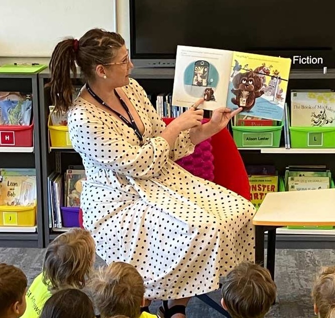 Sandy Robinson reading a picture book to a group of primary students in the College library, books and reading nooks visible behind her.